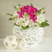 The image shows a compact yet luxurious flower arrangement that exudes a sense of calm celebration, the kind you might see on a beautifully laid table in a Stanmore home close to Canons Park. At its centre, a cluster of deep fuchsia blooms, reminiscent of full-bloom peonies, gathers tightly, each petal layered and softly ruffled. The colour is rich and inviting, somewhere between raspberry and plum, and the folds of the petals create a plush, cushion-like effect. Encircling this vivid core are graceful ivory alstroemeria, their petals long and slightly flared, kissed with fine streaks of pink and gold that resemble delicate brushstrokes. Between the flowers, small green leaves and upright buds add height, texture, and a sense of freshness, like new shoots appearing after a gentle rain. All of the blooms are nestled into a low, round white wicker basket, its interlaced bands flowing around the arrangement in neat, curved lines. The basket's bright whiteness and intricate weaving provide a chic, contemporary frame for the flowers' natural softness. Just in front of the basket sit two cream woven spheres, echoing the wicker texture and bringing an artistic, almost sculptural note to the scene. The light is soft and even, with no harsh shadows, allowing every petal edge and wicker strand to be seen clearly while maintaining a soothing atmosphere. This arrangement feels perfectly suited to wedding receptions, anniversaries, or milestone birthdays in Stanmore, and it's easy to imagine its subtle green, cool scent filling a room during a quiet family celebration or intimate dinner.
