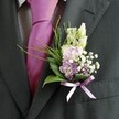 A close-up photograph shows the upper torso of a well-dressed man, most likely a groom or honoured guest at a Stanmore wedding, standing calmly in soft natural daylight. He wears a dark charcoal, almost black, tailored suit jacket, the texture of the fabric clearly visible along the notched lapel and sleeve. Beneath the jacket sits a crisp, pale dress shirt with a faint grid pattern, its tidy collar framing a bold magenta-fuchsia silk tie. The tie is carefully knotted and slightly lustrous, catching the light in gentle highlights that contrast with the matte suit cloth. Pinned neatly to the left lapel, just above the heart, is a meticulously crafted boutonnière. The floral arrangement combines slender, unopened white to creamy buds, reminiscent of snapdragons, rising gracefully at the top. They are cradled by clusters of soft pink and lavender-purple blooms-delicate sweet-pea-like flowers or stock-interwoven with deeper purple florets for subtle depth. Tiny cloud-like sprigs of white gypsophila nestle between the petals, adding a misty, romantic air. At the base, glossy dark green leaves form a supportive cradle, while finer, feathery bright green foliage fans outward with an airy, fern-like movement. A lavender satin ribbon, tied into a small, perfectly formed bow, secures the stems and echoes the lilac tones in the petals. The composition feels quietly celebratory, as if captured just outside a Stanmore ceremony venue or on a leafy street near Bentley Priory, and the careful coordination between tie and flowers suggests playful elegance, ideal for a local wedding morning or formal event portrait.