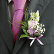 Pinned neatly against the dark, tailored fabric of a formal suit, this refined boutonnière offers a gentle contrast of colour and texture, much like a soft patch of wildflowers glimpsed along a quiet path near Bentley Priory in Stanmore. At its heart are lilac blossoms in a cool, pastel hue, their petals smooth and slightly curved, forming a small, layered cup that seems to hold the light. Nestled beside them, creamy white buds and open white flowers add brightness and purity, while tiny heads of crisp white baby's breath create a delicate, airy halo, softening the edges of the design. Around the blooms, a polished collar of deep green, curved garden leaves cradles the arrangement, their glossy surfaces reflecting subtle highlights, giving depth and structure. Slender blades of fresh green grass and fine, feathery shoots of foliage rise gracefully from the base, lending the piece a sense of height, movement, and natural spontaneity, as though it were freshly gathered from a spring border. The stems are carefully bound with a narrow lavender ribbon, tied so smoothly that the fabric lies flat and elegant, its muted sheen echoing the tones of the nearby tie and pocket square. The overall effect is one of understated charm and thoughtfulness, ideal for a wedding, prom, or special celebration in Stanmore's local venues. Created with the nuanced touch of a florist used to working on an intimate scale, this boutonnière suggests gentle fragrance, romantic sentiment, and the quiet pride of dressing well for a meaningful day.