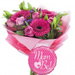 A joyful pink bouquet celebrating an expectant mother is shown in full detail against a clean white background, evoking the kind of warm, personal gift often sent across Stanmore for baby showers and pregnancy news. Rich fuchsia gerbera daisies form the main focus, their wide petals circling dark, velvety centres that create a striking contrast and a sense of depth. Between these bold blooms nestle soft pink roses, some tightly closed in bud, others half-open or nearing full bloom, their layered petals giving the arrangement a romantic, gently ruffled texture. Scattered throughout are sprays of tiny purple filler flowers, likely statice or limonium, adding dashes of lavender colour and a slightly papery delicacy that lightens the overall composition. Fresh green foliage frames the pinks, the leaves offering both structure and a cool, leafy backdrop that makes the flowers appear especially vibrant, as if freshly prepared in a Stanmore high-street studio just before delivery to a family celebration near Stanmore Hill. The bouquet is gathered into a rounded dome and carefully wrapped in layers of pink tissue paper, from bright fuchsia to pale pastel, forming a soft, voluminous collar around the stems. A layer of shiny clear cellophane hugs the outside, creating crisp edges and glints of reflected light that enhance the gift-like feeling. In front of the base of the bouquet sits a small, round, scalloped foil balloon, its bright pink surface dotted with white polka dots. In flowing white script is the heartfelt message Mum to Be!, with a small decorative heart-within-a-heart motif adding extra sweetness. The image conveys a mood of happiness, anticipation, and gentle celebration, perfectly suited to welcoming a new life in Stanmore.