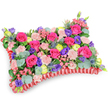 A vivid rectangular 'pillow' of flowers sits presented at a gentle oblique, slightly elevated angle against a clean white background, its surface a plush mosaic of colour and texture. At first glance the arrangement sings in hot fuchsia-pink roses, their velvety petals either tightly furled or just beginning to open, creating a soft, tactile rhythm. Interwoven between them are clusters of pale shell-pink carnations that ripple across the top like delicate folds of fabric. Deeper notes of purple lisianthus add bell-shaped contrast while cheerful lemon-yellow ranunculus pinprick the composition with sunny highlights. Cool, architectural flashes of silver-blue eryngium thistles introduce a spiky, wild element that contrasts beautifully with the rounded blooms. Sprays of silvery, rounded eucalyptus and darker green foliage nestle through the flowers, giving the pillow depth and a faint, herbaceous scent that balances the sweeter rose fragrance. The whole cushion is isolated by a rich, ruffled red satin ribbon base that hugs the perimeter, the pleated texture defining the classic pillow shape and lending a formal, memorial air. Soft studio light catches on the satin and the glossy petals, creating subtle shadows and a gentle sheen. Designed as a sympathy or memorial tribute, the palette - fuchsia, violet, lemon and blush - reads as a warm celebration of a life rather than austere mourning, offering comfort through colour and craft. As a Stanmore florist might note, this carefully balanced pillow would sit respectfully at a service or graveside, its lively tones and considered textures intended to console and recall fond memories in the quieter moments of remembrance.