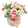 Centred against a spotless white background, a blush-pink cylindrical hatbox brims with an artfully arranged bouquet that looks freshly prepared in a Stanmore studio. The box's matte pastel surface provides a gentle contrast to the shine of the coral-red satin ribbon wrapped around its base and tied into a small, precise bow at the front. From this bow, a cream gift tag bearing elegant green script and a slender website line hangs lightly, suggesting a personalised floral delivery arranged with care. Above, the flowers rise in a rounded, dome-like shape that slightly overflows the rim, as if the blooms are leaning forward to greet the viewer. The focal point is a generous, fully open rose, its petals layered in a swirl of colour that moves from pale pink along the edges to a deeper, romantic pink at the centre. Around it, crisp white chrysanthemums or daisy-style blooms with bright green centres create a lively, structured ring. Threaded between are white alstroemeria, easily recognised by their tapered, trumpet-like petals accented with delicate speckles. Pops of deeper fuchsia or magenta from smaller flowers, perhaps spray roses or lisianthus, punctuate the softer tones and draw the eye across the bouquet. Eucalyptus leaves with rounded, silver-green shapes weave throughout, joined by darker green foliage and a scattering of tiny buds that bring a subtle, natural wildness to the design. The arrangement gives an impression of elegance without formality-perfect for same-day delivery around Stanmore, Canons Park, or Belmont as a birthday surprise, a romantic gesture, or a thoughtful way to say thank you in a style that feels both modern and warmly personal.
