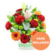 A joyful, colour-rich bouquet is arranged in a simple rectangular glass vase, wrapped with a light green and white gingham ribbon that lends the design an easy, welcoming charm, perfect for a home in Stanmore. The flowers are packed closely together, giving the impression of abundance and celebration. At the heart of the arrangement, deep red roses with velvety, tightly layered petals create a luxurious, romantic focal point. Around them, red and glowing red-orange carnations lend a soft, ruffled texture, their many petal layers making the bouquet appear generous and plush. Punctuating these warm tones are vivid orange gerbera daisies, each with a bold dark centre and a neat ring of flat petals radiating outward; they stand out like small suns, echoing the cheerful mood of a family gathering near Stanmore Station. Between these showy blooms, rounded clusters of lime-green button chrysanthemums nestle like bright beads, adding an invigorating pop of green that connects visually to the ribbon on the vase. Rising elegantly through the bouquet, white Oriental lilies bring height and grace, some flowers fully open to reveal gentle pink stripes and dustings of pollen, others still closed as slender buds, suggesting that the arrangement will continue to change and surprise over time. A mix of broad, glossy dark green leaves and slender, silver-green eucalyptus stems curls around the flowers, framing them and adding both depth and a cool, slightly woodland note to the overall effect. The stems, visible through the clear water in the vase, emphasize the bouquet's freshness and clean lines. In the lower right corner of the image, an orange circular badge with bold white text announces VASE INCLUDED, clearly indicating that this bright, uplifting display arrives ready to place on a table, desk, or mantelpiece anywhere in Stanmore.