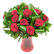 A lush, rounded bouquet of a dozen or more deep crimson roses fills the frame, each bloom at peak openness with velvety, wine-dark petals spiraling from a tight centre to reveal layered texture and a fresh, fragrant heart. The roses are nestled in a generous collar of dark green foliage-glossy, flat leaves like salal or ruscus form a dense backdrop while broader, emerald aspidistra leaves are expertly folded and rolled into sculpted loops and spirals that cradle the heads and introduce an architectural, contemporary rhythm. Those curved green ribbons catch light differently from the soft petals, creating a tactile contrast between the satiny reds and the sleek, almost lacquered greens. The stems sit in a tall, translucent ruby-red glass vase with a subtle ribbed or twisted surface; its flared mouth, narrowed waist and stable base create elegant vertical lines that echo the roses' stems and catch reflections of light to intensify the bouquet's passionate tones. The arrangement sits against a pure white background under soft, even lighting, which sharpens the saturated reds and the deep greens while allowing every petal edge and leaf fold to read clearly to the eye. The overall effect is classic romance with a modern twist-perfect as an anniversary or Valentine's centrepiece or as a heartfelt gift that evokes formal elegance and warm intimacy. As a local florist in Stanmore I picture this on a candlelit table near Stanmore High Street or sent to a loved one after a stroll through Bentley Priory, where the scent and colour speak as eloquently as words.