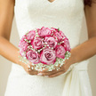 The image presents a close-up of a bridal bouquet being cradled softly against an ivory lace wedding dress, a familiar scene for ceremonies taking place in Stanmore churches and venues near Canons Drive. The bouquet itself is compact and impeccably shaped, composed almost entirely of blush-pink roses arranged into a neat, rounded cluster. Each rose is at full bloom, petals overlapping in tight yet fluffy layers that give the bouquet a plush, cushion-like appearance. The colour palette remains restrained and romantic: gentle blush tones with hints of cream and warm pink blending seamlessly from one bloom to the next. Interspersed among the roses are fine sprays of white baby's breath, known as gypsophila, whose tiny clustered blossoms look like delicate flakes of snow scattered through the bouquet. These light, cloud-like accents soften the lines where the roses meet and add an ethereal touch, creating a sense of air and lightness that contrasts beautifully with the solid, velvety rose heads. The bride's hands hold the stems just beneath the blooms, fingers relaxed, suggesting both composure and quiet anticipation as though she might be standing just outside a reception at Bentley Priory Museum or near Stanmore Country Park. Her lace gown forms an elegant backdrop, its floral motifs and sheer texture subtly mirroring the bouquet's romantic character. Soft ambient light, perhaps from an overcast North London sky, washes over the arrangement and the fabric, deepening the pastel tones and revealing fine details in both petal and lace without harsh contrasts. You can almost imagine the fragrance rising gently: a clean, fresh rose scent with a hint of greenery, soothing and intimate. The overall mood is classic and deeply romantic, a bouquet designed to accompany a Stanmore bride through every cherished moment of her wedding day, from walking down the aisle to the final dance.