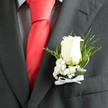 In soft, even light the ivory rose boutonnière takes centre stage against a smart charcoal jacket, the bloom's velvety petals arranged to show a gentle spiral and an open heart. Around it, frothy sprays of baby's breath scatter tiny points of cream that diffuse the rose's presence into something tender and luminous. Slender blades of dark green foliage arch and curl, their glossy surfaces providing contrast and a cool, natural frame; fine, twisted leaves add a whisper of movement that catches when the wearer moves. The boutonnière sits beside the crisp edge of a checked shirt and the confident colour of a red silk tie, creating a classic, slightly romantic pairing that reads as both timeless and personal. A neat white ribbon bow at the base ties the stems together, the satin reflecting the light like a small pledge of care. You can almost sense the rose's subtle fragrance - a cool, green perfume that speaks of early-morning dew and careful handling. As local florists preparing arrangements for Stanmore and Canons Park know well, this composition is chosen to harmonise with formal wear while remaining quietly expressive: suitable for grooms, fathers of the bride, or anyone whose day calls for a single, meaningful flourish. Flower Delivery Stanmore often composes pieces like this for weddings on Stanmore High Street and intimate ceremonies near the tube station, where attention to texture and freshness matters as much as appearance.