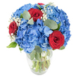 A lush, sculpted bouquet sits in a simple clear glass vase, photographed against a crisp white backdrop so every shade and texture reads clearly. The arrangement is anchored by voluminous clusters of sky-blue hydrangeas whose many small florets form a soft, cloud-like dome; the blue is bright and slightly cool, as if recalling a summer sky after rainfall. Punctuating that sea of blue are three deep, velvety red roses with tightly furled petals - concentrated points of passionate colour that draw the eye inward. Sprigs of tiny white baby's breath (Gypsophila) float between the larger blooms like delicate snowflakes, their fine stems adding an airy lightness and gentle contrast. Rounded, muted-green eucalyptus leaves are woven through the composition, their smooth, silvery-green surfaces and subtle herbal scent lending calm depth and an organic silhouette. The vase's gently rounded base and tapered mouth reveal crisp green stems submerged in clear water, emphasising the bouquet's freshness while the bright, natural lighting softens shadows and highlights petal texture. The overall mood is optimistic and quietly celebratory - appropriate for anniversaries or a birthday, an intimate romantic gift, or a striking centrepiece for a dining table at home. As a local Stanmore florist might note, this is the kind of arrangement you could imagine placed on a mantel after a walk on Stanmore Common or sent to a neighbour on Stanmore High Street to brighten their day; it looks as comfortable in a modern flat as it does in a traditional bay-windowed cottage. The colours together - clear blue, passionate red, and tender white - feel both fresh and timeless, offering warmth, refinement and a gentle aromatic lift from the eucalyptus.