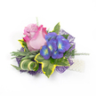 Pinned against a crisp white backdrop, this compact corsage reads like a tiny, wearable garden. At its heart sits a single partially open rose whose petals graduate from a soft lavender-pink at the centre to a brighter fuchsia blush at the outer edges, offering a velvet softness you can almost smell - a gentle, fresh scent that hints at morning flowers. To its right, a small cluster of indigo-purple florets, reminiscent of hydrangea or delicate orchids, introduces jewel-toned contrast with tiny bright blue and lime-yellow-green centres that catch the light like garden jewels. Surrounding the blooms is a thoughtful assortment of foliage: broad variegated ivy leaves with deep emerald cores and sunlight-yellow margins, slender silvery-green leaves that suggest lavender or rosemary foliage, and airy, wispy asparagus-fern tendrils that give the piece breathing room. A curled, striped green-and-cream leaf provides a sculptural twist, while a foundation of deep purple, web-like mesh peeks out beneath the arrangement, adding contemporary depth and a soft shimmer. A short strand of luminous white pearls emerges at the lower right, implying a wristlet or lapel attachment and lending a timeless touch. The composition is lit by soft, even daylight that reveals texture and layered depth without harsh shadows, and it rests cleanly on white to keep focus on colour and form. Ideal for weddings, proms or milestone celebrations, this corsage balances romantic blooms with modern accents, evoking quiet joy and careful craftsmanship by a florist who values both tradition and contemporary detail.