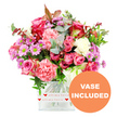 A lively and affectionate floral arrangement fills a clear glass cube vase, photographed on a bright white background that accentuates every shade and texture. The bouquet is dominated by pink tones: large roses in deep fuchsia and soft blush open fully at the top, their petals layered in satin-like whorls that immediately convey romance. Around them, generous carnations in coral, peach-pink, and creamy white add density and softness, their ruffled petals creating a plush, almost cushion-like effect. Nestled between these larger blooms are small daisy-like chrysanthemums in light purple and lavender, their vivid green centres glowing like tiny jewels that bring a playful twist to the palette. The flowers are enveloped in a mix of foliage: rounded eucalyptus leaves in a cool, silvery-green, darker glossy leaves that anchor the design, and a few hints of reddish-brown foliage that introduce warmth and depth, reminiscent of the rich tones seen around Stanmore's parks in late afternoon light. The stems are neatly aligned and clearly visible through the square vase, which has smooth, transparent sides that lend a contemporary edge. Around the lower portion of the vase, a white decorative band wraps neatly, printed with small red hearts and the tender phrase all my love in a cursive red script, turning the base into a visual love note. In the lower right corner of the photograph, a bold orange circular graphic with clean white capital letters states VASE INCLUDED, making it clear that this is a ready-to-display gift, ideal for setting straight onto a desk or table in any Stanmore home or office. The overall mood is joyful, loving, and perfectly suited to birthdays, anniversaries, Mother's Day, or any occasion when you want your feelings to be seen as well as felt.