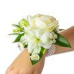 A delicate wrist corsage captured against a clean white background: at its heart sits a creamy ivory rose, petals unfurling with a velvety softness that catches a cool, diffused light and lends the piece a gentle, luminous centre. Nestled just below and slightly to the left, a larger white trumpet-shaped bloom - reminiscent of a Peruvian lily or a small trumpet lily - opens to reveal a soft green-yellow throat that gives the arrangement depth and a whisper of spring. Small, vibrant green buds peek between the main flowers, hinting at future blooms, while airy sprays of Baby's Breath (Gypsophila) weave a cloud-like texture around the composition. Near the wrist, a cluster of tiny daisy-like white florets with bright yellow centres adds a playful, sunlit detail. Broad, glossy deep-green leaves frame the flowers, their smooth, waxy surfaces creating a lush contrast to the pristine whites. The whole ensemble is secured by a chic multi-strand pearl bracelet: lustrous, uniformly round pearls loop gently around the wrist, the cool smoothness of the beads balancing the tender floral softness. Imagine the faint, clean scent of petals and green stems, a powdery, comforting aroma that evokes early spring mornings in Stanmore near Canons Park. Crafted with careful wiring and thoughtful placement, this corsage reads as both timeless and intimate - an accessory suited to weddings, proms, or any formal celebration where a quiet, refined statement is wanted. As a local florist might explain to a customer arranging delivery across Stanmore High Street and nearby streets, the piece is designed to look effortless on the wrist while remaining secure and freshly vibrant through an evening of vows, dances, and photographs.